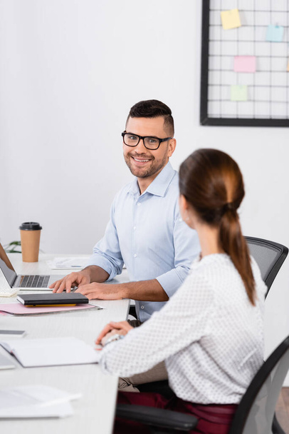 cheerful businessman in glasses looking at businesswoman sitting at desk on blurred foreground - Foto, imagen
