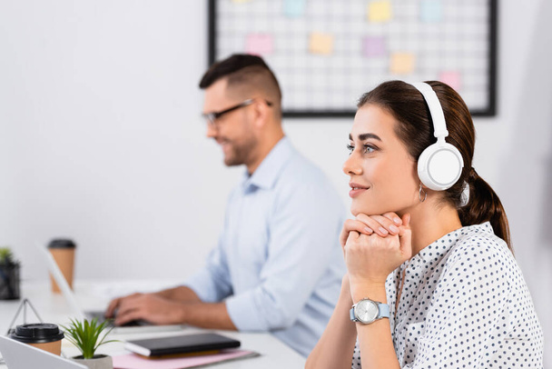 thoughtful businesswoman in wireless headphones near businessman working on blurred foreground - Фото, изображение
