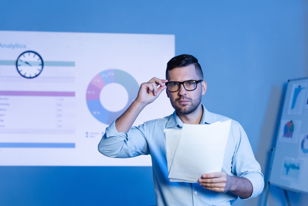 businessman adjusting glasses and holding papers near charts and graphs on wall - Foto, imagen