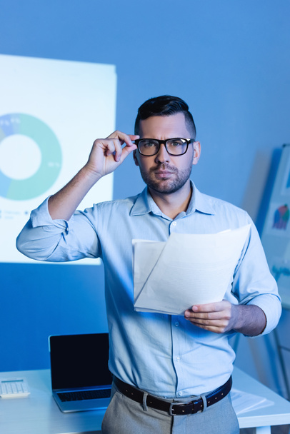 businessman adjusting glasses and holding documents near charts and graphs on wall - Foto, Imagen