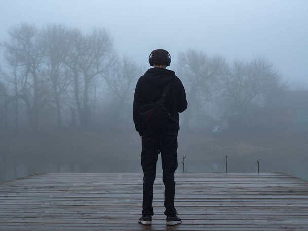 A young man stands alone on a wooden bridge by the lake. Early cloudy foggy morning. - Photo, Image
