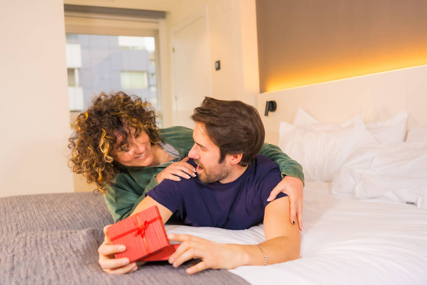 Valentine's Day, to a young Caucasian couple in pajamas on the bed, the bride giving a nice surprise to the boyfriend with a gift. With copy and paste space on the right - Foto, Imagen