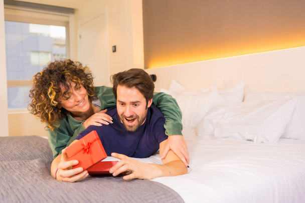 Valentine's Day, to a young Caucasian couple in pajamas on the bed, the bride giving a nice surprise to the boyfriend with a gift. With copy and paste space on the right - Valokuva, kuva
