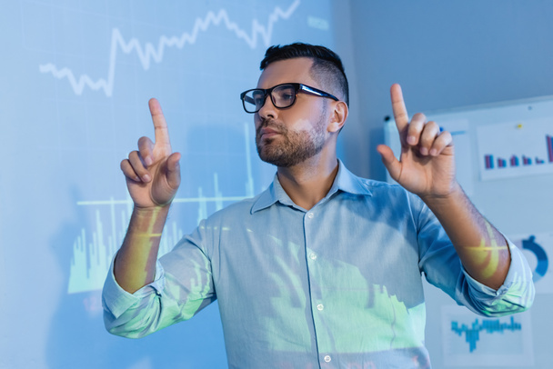 businessman in glasses pointing with fingers near digital graphs in office - 写真・画像