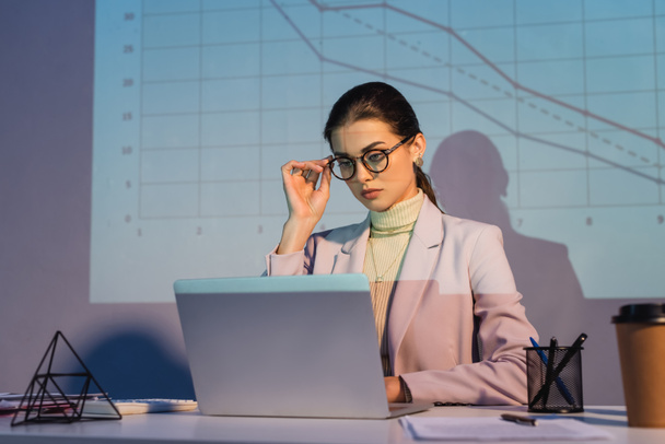 mujer de negocios ajuste de gafas y el uso de ordenador portátil cerca de gráficos digitales en la pared y taza de papel en primer plano borrosa - Foto, imagen