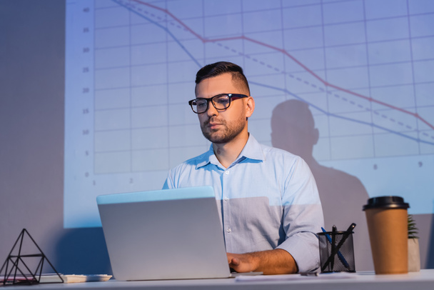businessman in glasses looking at laptop near graphs on wall and paper cup on desk - Foto, Imagen