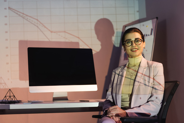 happy businesswoman in glasses looking at camera while sitting near computer monitor with blank screen - Zdjęcie, obraz
