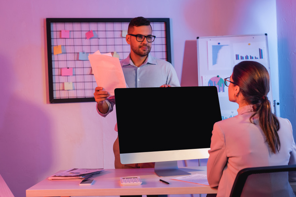 empresaria sentada cerca de monitor de computadora con pantalla en blanco y mirando a compañero de trabajo con documentos  - Foto, Imagen