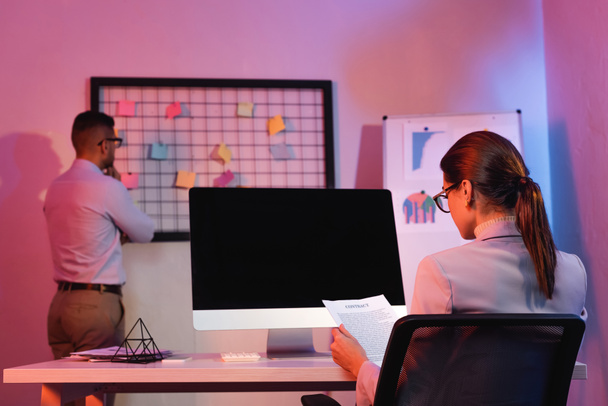 İş kadını boş ekran ile bilgisayar monitörünün yanında oturuyor ve bulanık arkaplan üzerinde iş arkadaşı sözleşmesine bakıyor - Fotoğraf, Görsel