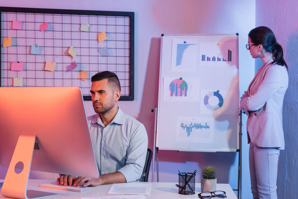 επιχειρηματίας κοιτάζοντας οθόνη υπολογιστή, ενώ επιχειρηματίας στέκεται κοντά flipchart σε θολή φόντο - Φωτογραφία, εικόνα
