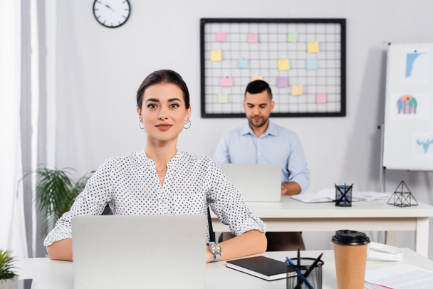 cheerful businesswoman smiling near laptop on desk and coworker on blurred background  - Foto, Bild