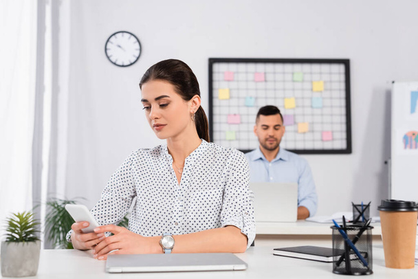 businesswoman using smartphone near laptop on desk and coworker on blurred background  - Zdjęcie, obraz
