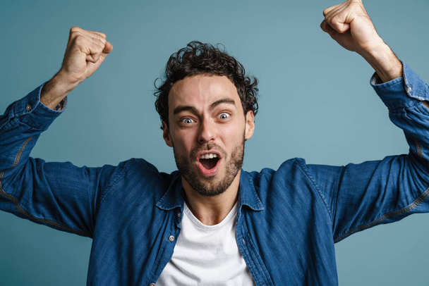 Excited caucasian guy screaming and making winner gesture isolated over blue background - Foto, afbeelding