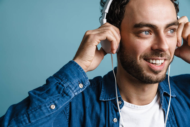 Smiling handsome guy listening music with headphones isolated over blue background - Fotó, kép