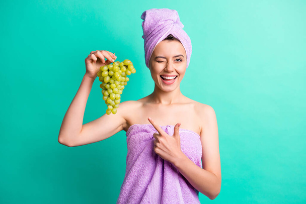 Photo of funny girl indicate finger grape bunch close wink eye wear violet towel turban isolated teal color background - Foto, afbeelding