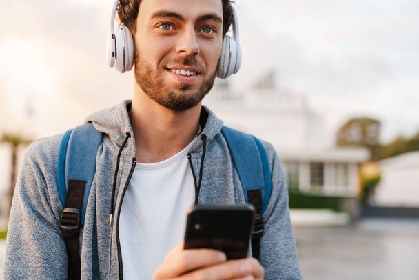 Smiling young guy in headphones using mobile phone while walking on promenade - Fotó, kép