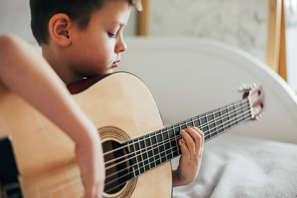 Boy 6-7 years old boy playing guitar indoor. Hobby leisure concept. Authentic lifestyle moment - Photo, Image