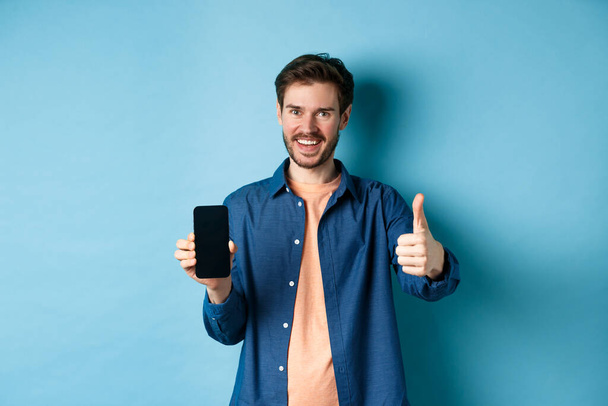 Handsome young man showing thumbs up gesture and empty smartphone screen, recommending app or company, standing on blue background - Фото, изображение