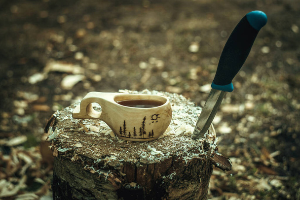 Ein Holzbecher aus Holz mit schwarzem Tee, neben einem Messer, das im Holz steckt. Zeichnung des Waldes auf dem Kreis. - Foto, Bild