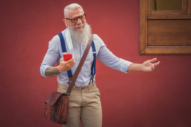 Пожилой мужчина в хипстерской одежде, очках и длинной белой бороде ходит и разговаривает по телефону, сосредоточившись на голове - Фото, изображение