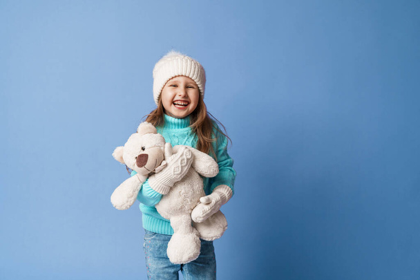 happy little 5-year-old girl with blonde hair in a knitted sweater cap and mittens, smiles and hugs a teddy bear on a blue background in the studio. The toy is her friend. Winter time. Holiday weekend - Foto, imagen