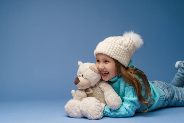 happy little 5-year-old girl with blonde hair in a knitted sweater cap and mittens, smiles and hugs teddy bear lying on blue background in the studio. toy is her friend. Winter time. Holiday weekend - Фото, изображение