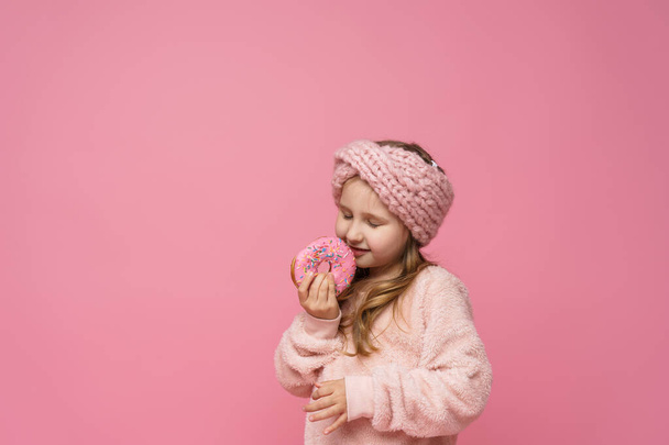 little girl in a fluffy sweater and scarf, smiling with a doughnut in her hand. Sweet appetizer, delicious pastries. a child shows a delicious dessert on a pink background in studio - Photo, Image