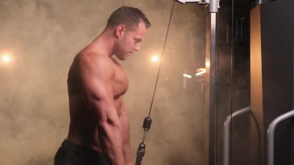 Hombre fuerte haciendo ejercicio con la moderna máquina de entrenamiento en el gimnasio brillante y ahumado - Imágenes, Vídeo