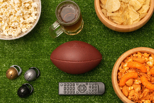 Super bowl flat lay photography with snacks, beer, american football ball, little helmets and remote control on green grass background - Foto, Bild