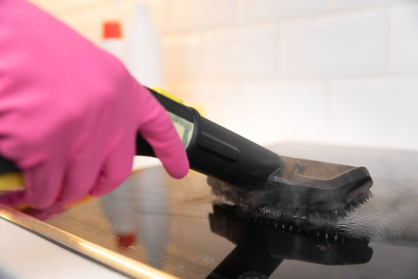 Femme au foyer en gants de caoutchouc nettoyage table de cuisson en céramique noire avec un nettoyeur à vapeur chaude. Nettoyage du printemps. - Photo, image