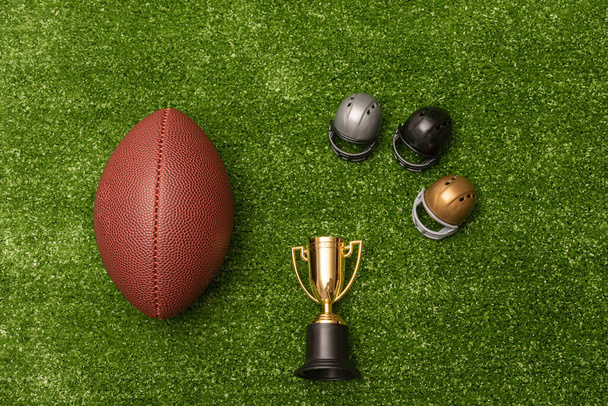 Американский футбольный мяч, маленькие футбольные шлемы и золотой трофей на зеленом фоне - Фото, изображение