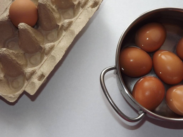 béžová vejce vařená v hrnci vody s krabicí od vajíček na šedém pozadí - Fotografie, Obrázek
