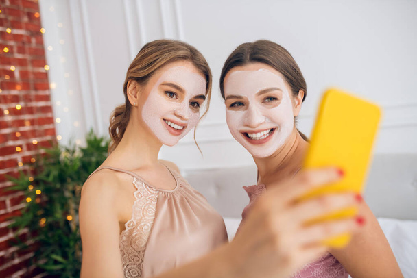 Two cute girls with facial masks making selfie - Foto, imagen