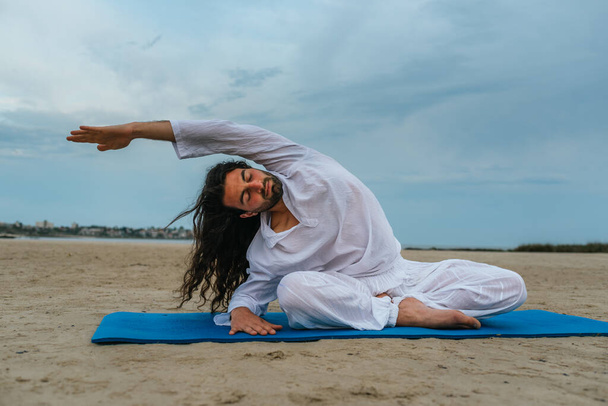 Foto de stock de hombre adulto con el pelo largo haciendo yoga posa en la playa. - Foto, imagen