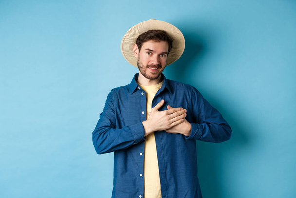 Handsome traveler in straw hat, holding hands on heart and smiling, showing thank you gesture, keep memories about vacation in soul, standing on blue background - Foto, imagen