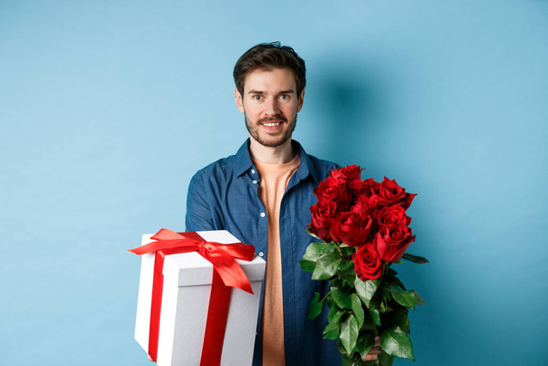 Láska a Valentýn koncept. Okouzlující mladý muž dává dárek a kytice růží na přítelkyni, stojící nad modrým pozadím - Fotografie, Obrázek