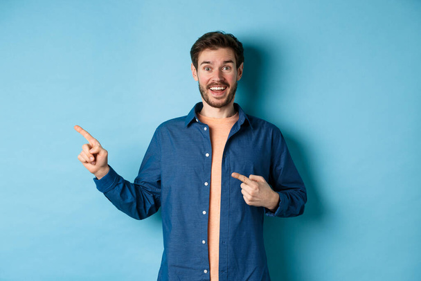 Podekscytowany i szczęśliwy biały mężczyzna uśmiecha się do kamery, wytykając palcami w lewo pustą przestrzeń, pokazując logo, stojąc na niebieskim tle - Zdjęcie, obraz