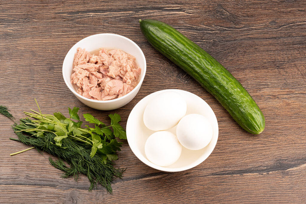 Fresh cucumber, parsley with dill, tuna and white eggs on a wooden background. Salad ingredients. - Photo, Image