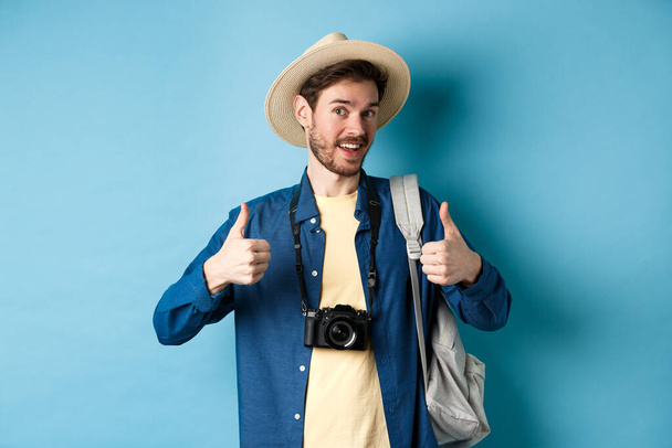 Happy smiling tourist say yes and show thumbs up, going on summer vacation, holding backpack and photo camera, blue background - 写真・画像