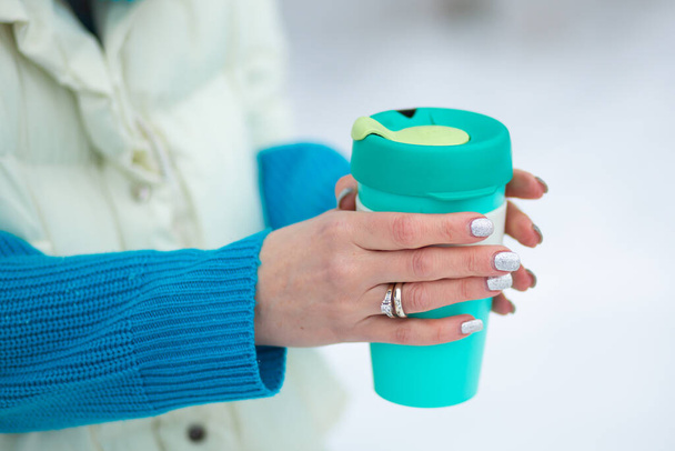 Les mains de la femme tenant une tasse thermique verte dans une journée d'hiver avec une neige blanche sur un fond. - Photo, image