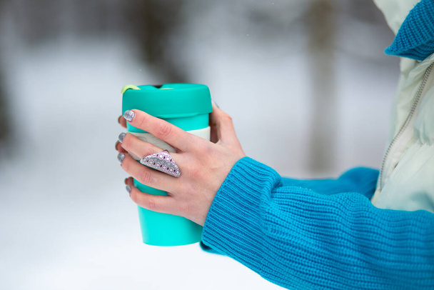 Les mains de la femme tenant une tasse thermique verte dans une journée d'hiver avec une neige blanche sur un fond. - Photo, image