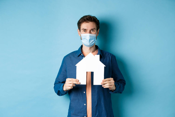 Emlak ve karantina konsepti. Tıbbi maskeli mutlu genç adam ev kesimini gösteriyor, mülk alıyor, mavi arka planda duruyor. - Fotoğraf, Görsel