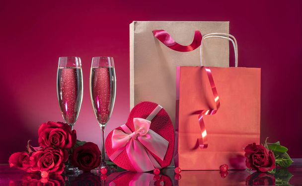Şatafatlı kadehler, köpüklü şarap ve saten yaylı bir kalp şeklinde hediye kutusu ve kırmızı arka planda ayna yüzeyinde güllerle çevrili bir alışveriş paketi. Sevgililer Günü ya da romantik akşam daveti - Fotoğraf, Görsel