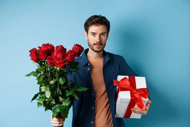 Romance and valentines day. Man presenting bouquet of red roses to lover. Boyfriend bring flowers and gift on romantic date, standing over blue background - Φωτογραφία, εικόνα