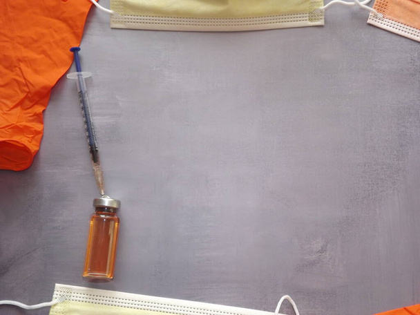 sobre un fondo gris, una vacuna, una jeringa, máscaras y guantes médicos de color naranja - Foto, Imagen