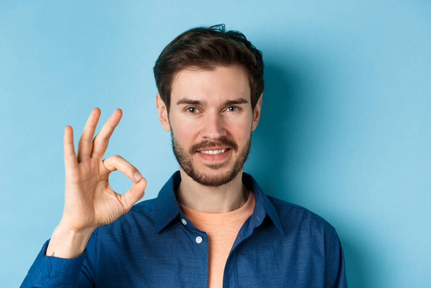 Detailní záběr pohledný vousatý chlap ukazuje v pořádku gesto a usmívá se, stojí na modrém pozadí - Fotografie, Obrázek