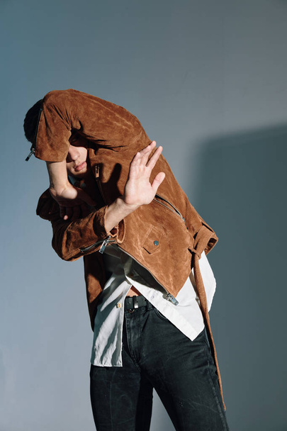 A man in a brown jacket covered himself with his hands from the light on a gray background - Foto, immagini