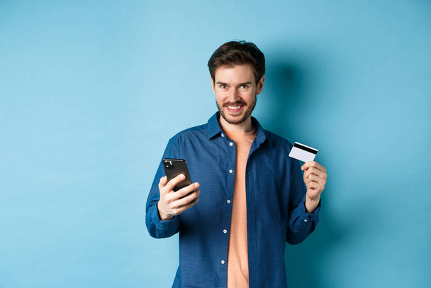 Koncept elektronického obchodu. Fešák mladý muž nakupování on-line, drží smartphone a plastové kreditní karty, usmívá se na kameru, stojí na modrém pozadí - Fotografie, Obrázek