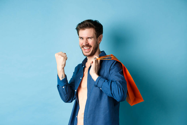 Vrolijke man die ja zegt, vuistpomp omhoog en glimlacht, oranje boodschappentas vasthoudt, vreugde voelt na het kopen met kortingen, op een blauwe achtergrond staat - Foto, afbeelding