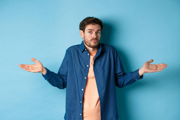 Confused young man with beard, shrugging shoulders and looking clueless at camera, know nothing, standing on blue background - Photo, image